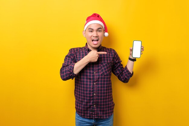 Sorpreso giovane uomo asiatico in cappello di Babbo Natale che punta le dita sullo smartphone con schermo vuoto su sfondo giallo dello studio Felice anno nuovo 2023 celebrazione concetto di vacanza allegra