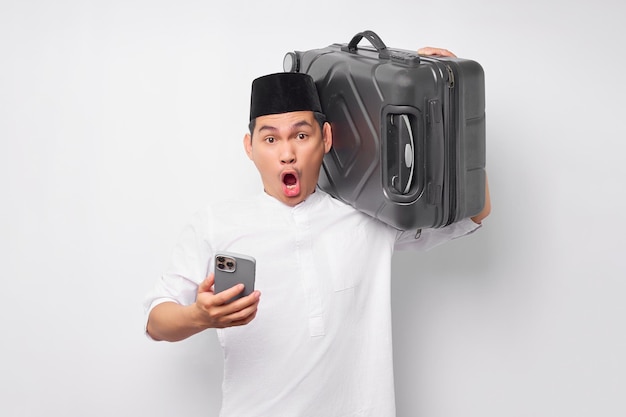 Sorpreso giovane uomo musulmano asiatico che porta una valigia e utilizza il telefono cellulare mentre reagisce alle cattive notizie isolate su sfondo bianco Ramadan e concetto di eid Mubarak