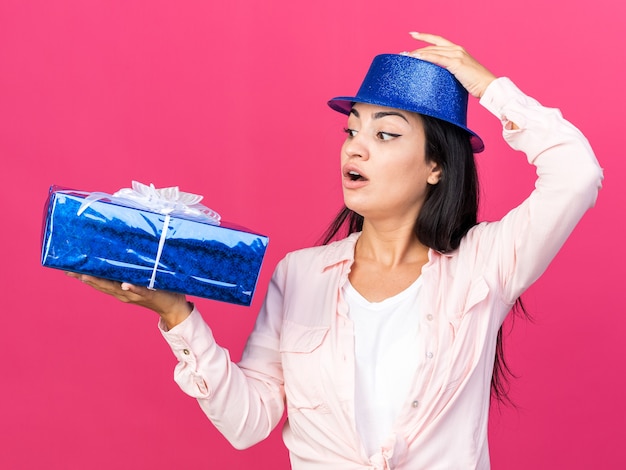 Sorpreso lato giovane bella ragazza che indossa un cappello da festa con scatola regalo