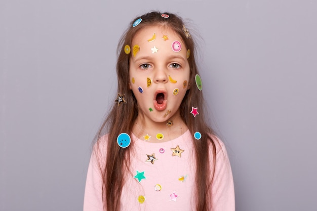 Sorpreso scioccato bambina coperto adesivi colorati per bambini in piedi isolato su sfondo grigio guardando la telecamera dicendo wow vede qualcosa di sorprendente