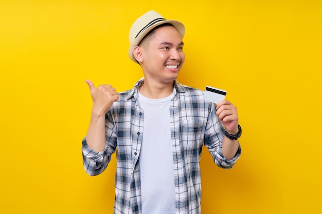 Sorridendo felice etnia asiatica giovane uomo 20s indossando abiti casual cappello tenendo carta di credito in mano pollice puntato da parte isolato su sfondo giallo ritratto in studio persone concetto di stile di vita