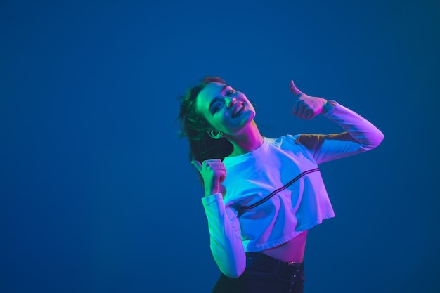 Sorridendo, pollici in su. Ritratto di giovane donna caucasica isolato su sfondo blu studio in luce al neon. Bellissimo modello femminile. Concetto di emozioni umane, espressione facciale, vendite, pubblicità, cultura giovanile.