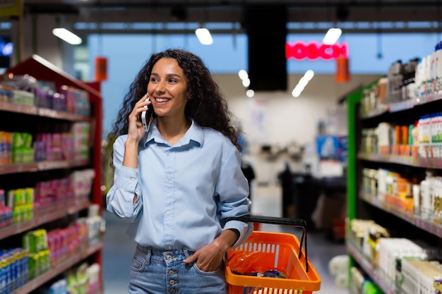 Sorridente acquirente donna ispanica in supermercato cammina tra file di scaffali con merci donna