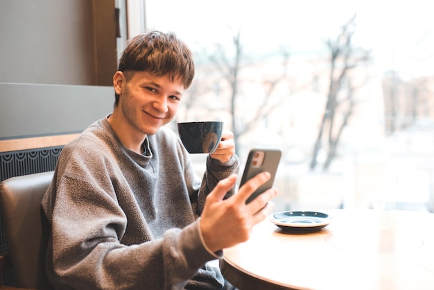 Sorridente adolescente gen Z ragazzo di 17-18 anni che beve caffè e tiene il telefono cellulare seduto nella caffetteria. Felice uomo millenario chat con gli amici con una tazza di tè divertirsi. Posto di lavoro.