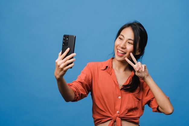 Sorridente adorabile femmina asiatica che fa foto selfie su smart phone con espressione positiva in abbigliamento casual e stand isolato sulla parete blu. La donna felice adorabile felice si rallegra del successo.