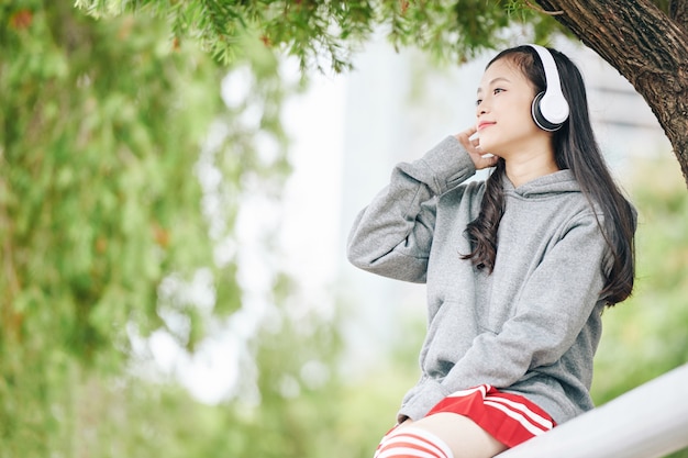 Sorridente affascinante giovane ragazza vietnamita che trascorre del tempo nel parco e ascolta musica in cuffia