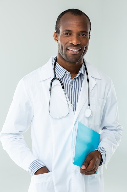 Sorridente afro americano medico in piedi contro il muro bianco mentre si tiene un caso clinico