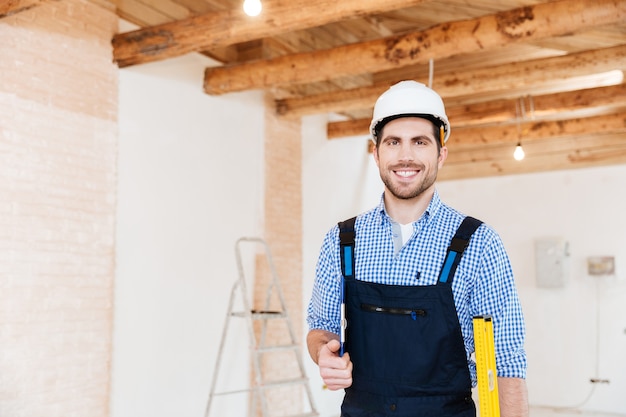 Sorridente allegro costruttore in piedi e tenendo gli strumenti sul posto di lavoro al chiuso