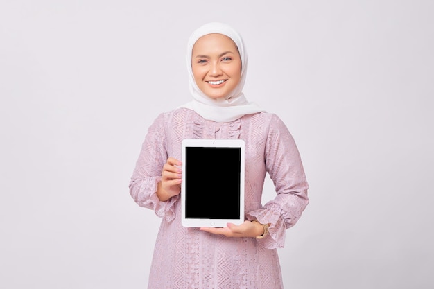 Sorridente amichevole giovane donna musulmana asiatica che indossa hijab e abito viola che mostra tablet digitale con schermo vuoto per mockup isolato su sfondo bianco studio