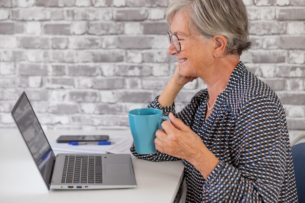 Sorridente anziana anziana donna d'affari seduta alla scrivania in videoconferenza con il computer portatile che tiene una tazza di caffè blu