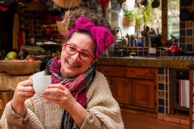 Sorridente anziana donna latina seduta in una cucina che indossa una sciarpa in testa mentre beve il caffè