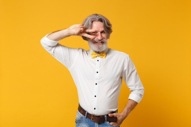 Sorridente anziano uomo con baffi dai capelli grigi barbuto in camicia bianca e cravatta a farfalla in posa isolata su sfondo giallo arancione in studio. Concetto di stile di vita delle persone. Mock up spazio di copia. Mostra il segno della vittoria.