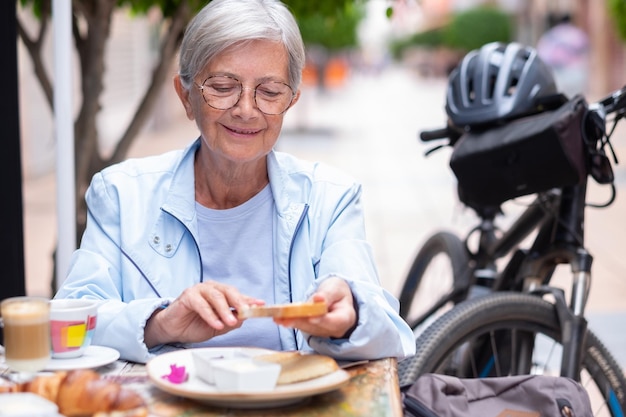 Sorridente attraente donna dai capelli grigi che tiene una fetta di pane tostato mettendo la marmellata sopra mentre si siede al tavolo di un bar all'aperto per colazione Donna anziana vicino alla sua bicicletta elettrica godendosi un momento di relax