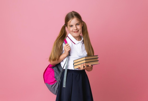 Sorridente bambina bionda 1012 anni in uniforme scolastica con zaino in possesso di libri isolati su sfondo rosa pastello ritratto in studio Il concetto di stile di vita dei bambini Istruzione a scuola