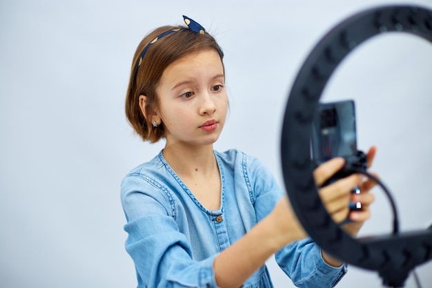 Sorridente bambina blogger in abito casual in denim usa selfie lampada a led e smartphone