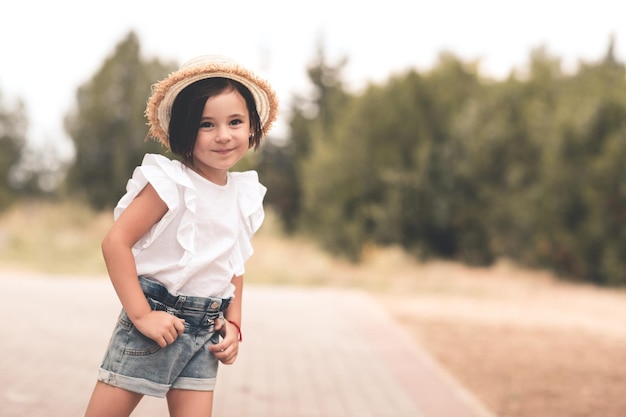 Sorridente bambina che indossa cappello di paglia top bianco e pantaloncini di jeans