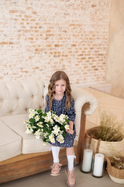 Sorridente bambina riccia con bouquet di fiori di gelsomino si siede sul divano in soggiorno Celebration