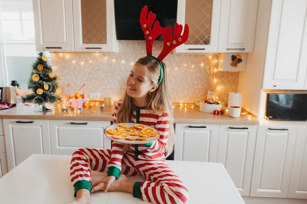 Sorridente bambina seduta al tavolo bianco in cucina con le arance. Giochi per bambini nella cucina addobbata per Natale. Bambina felice alla vigilia di Natale a casa