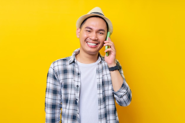 Sorridente bel giovane asiatico 20 anni che indossa abiti casual cappello parlare parlando al cellulare cellulare conducendo una piacevole conversazione isolata su sfondo giallo Concetto di stile di vita delle persone