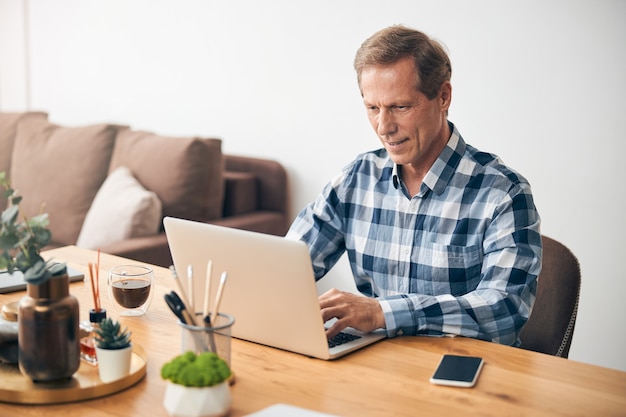 Sorridente bell'uomo adulto che esprime positività mentre legge i messaggi sul laptop a casa