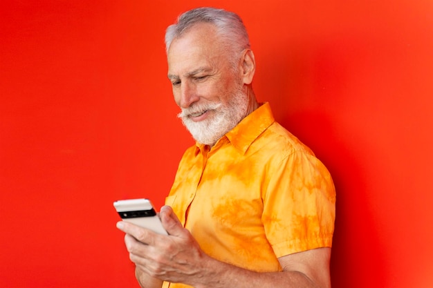 Sorridente bell'uomo anziano che tiene il telefono cellulare leggendo messaggi di testo shopping online isolato su sfondo rosso Hipster barbuto utilizzando la comunicazione smartphone sulla strada Concetto di mobile banking