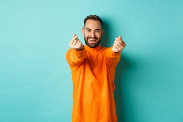 Sorridente bell'uomo che mostra i cuori e sembra felice, gesto fortunato, in piedi con un maglione arancione su sfondo azzurro