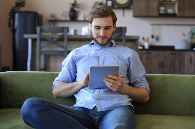 Sorridente bell'uomo che tiene e usa tablet con i social network a casa, felice libero professionista che fa shopping online, seduto su un comodo divano in soggiorno.