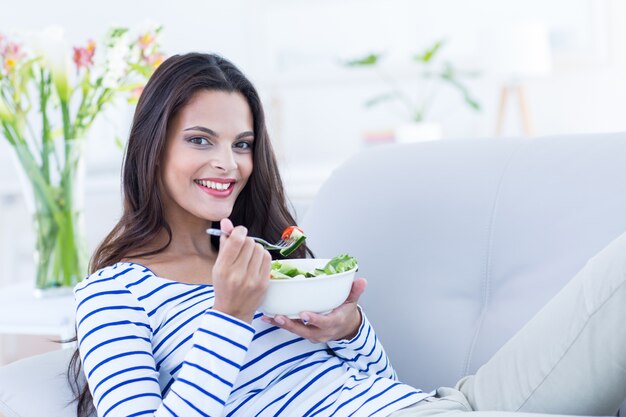 Sorridente bella bruna rilassante sul divano e mangiare insalata