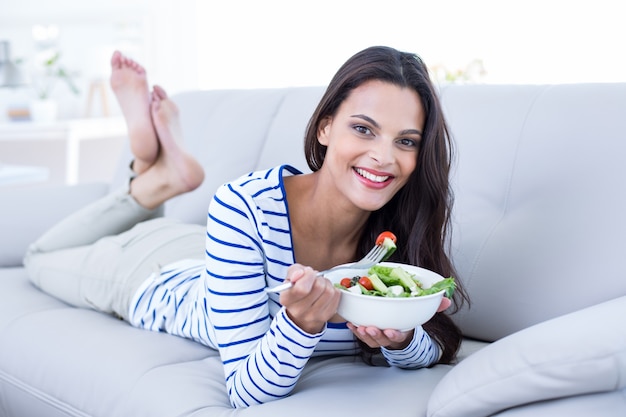 Sorridente bella bruna rilassante sul divano e mangiare insalata