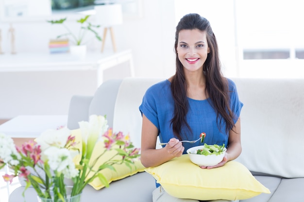 Sorridente bella bruna seduta sul divano e mangiare insalata