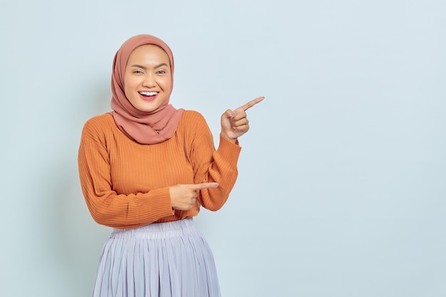 Sorridente bella donna asiatica in maglione marrone e hijab che punta il dito nello spazio della copia che mostra i prodotti pubblicitari e guardando fiducioso la fotocamera isolata su sfondo bianco