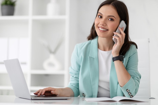 Sorridente bella donna caucasica millenaria manager in abito digitare su computer chiamate per telefono godere