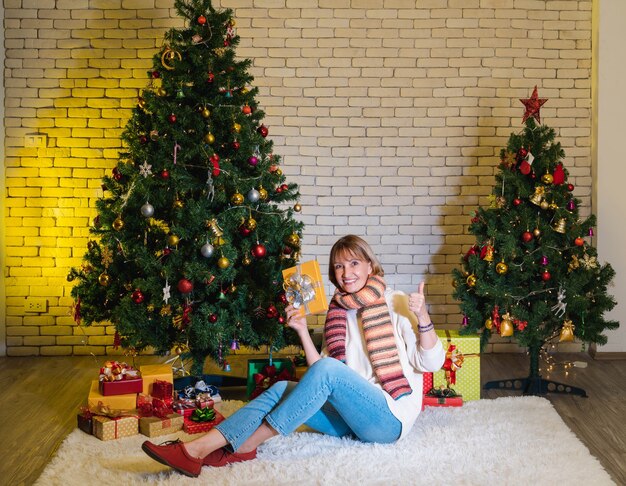 Sorridente bella donna caucasica senior azienda regalo di Natale e pollice in su. donna felice si siede davanti a un albero di Natale decorato con regali.