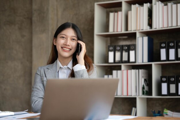 Sorridente bella donna d'affari asiatica che analizza grafico e grafico che mostra i cambiamenti sul mercato e tiene lo smartphone in ufficio