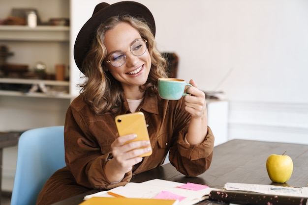 sorridente bella donna in occhiali e cappello usando il cellulare mentre beve il caffè a casa