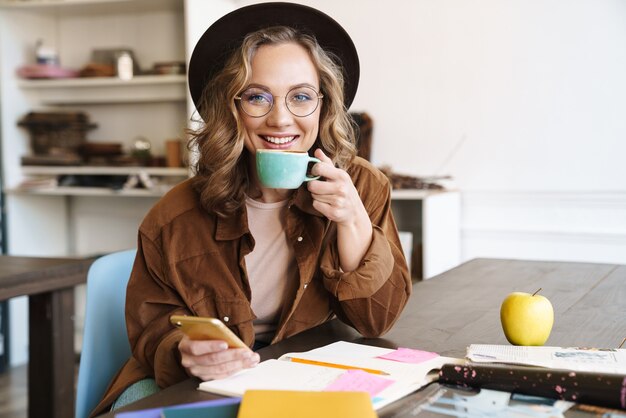 sorridente bella donna in occhiali e cappello usando il cellulare mentre beve il caffè a casa