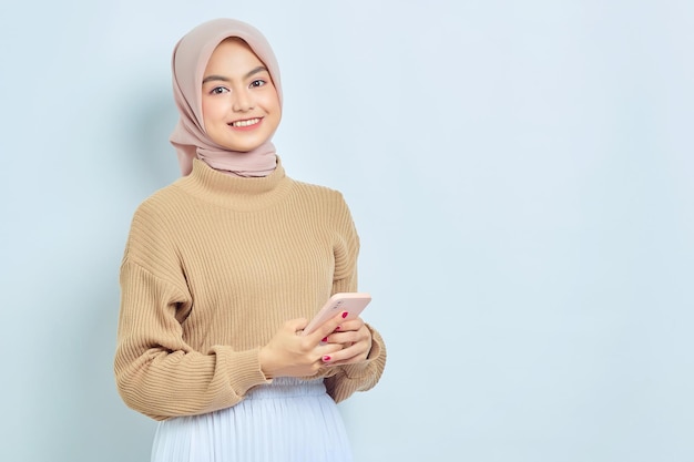 Sorridente bella donna musulmana asiatica in maglione marrone e hijab utilizzando un telefono cellulare ha ricevuto buone notizie isolate su sfondo bianco Persone concetto di stile di vita religioso