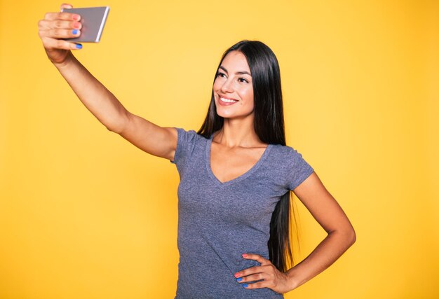 Sorridente bella e carina giovane donna fa selfie sul suo moderno smartphone isolato su muro giallo Foto sulla memoria Immagine sul telefono cellulare