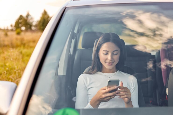 Foto sorridente bella giovane donna adulta guardando il suo telefono cellulare mentre è seduto all'interno dell'auto attraente donna dai capelli scuri che indossa una camicia bianca nella sua automobile che naviga in internet tramite smart phone