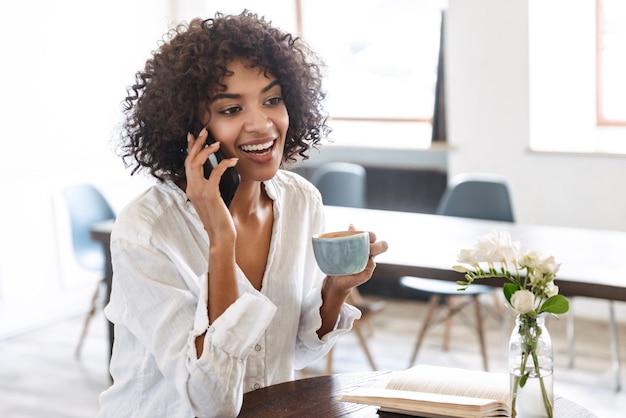 Sorridente bella giovane donna africana rilassante al chiuso, utilizzando il telefono cellulare, parlando