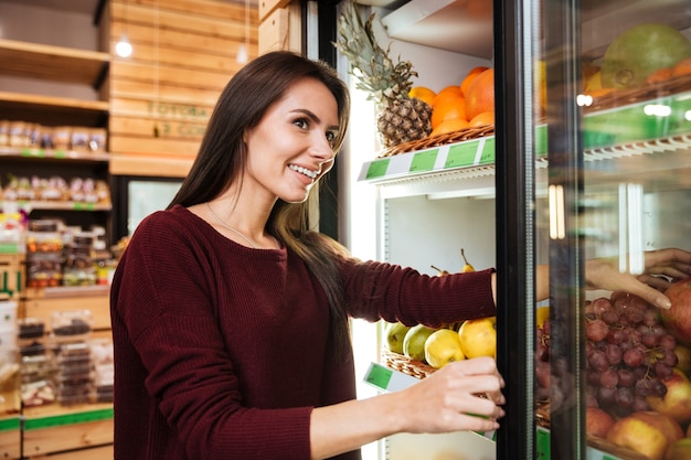 Sorridente bella giovane donna che sceglie e compra frutta al negozio di alimentari