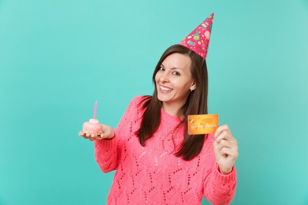 Sorridente bella giovane donna in maglione rosa lavorato a maglia, cappello di compleanno tenere in mano torta con candela, carta di credito isolata su sfondo blu muro turchese. Concetto di stile di vita della gente. Mock up copia spazio.