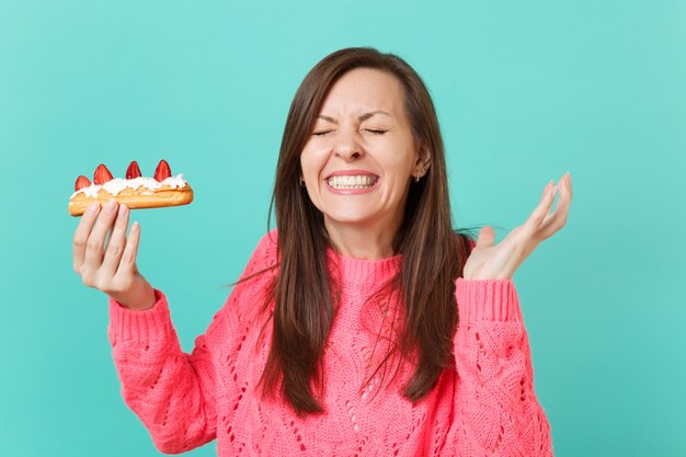 Sorridente bella giovane donna in maglione rosa lavorato a maglia con gli occhi chiusi che allargano le mani, tenere in mano la torta eclair isolata su sfondo blu ritratto in studio. Concetto di stile di vita della gente. Mock up copia spazio.