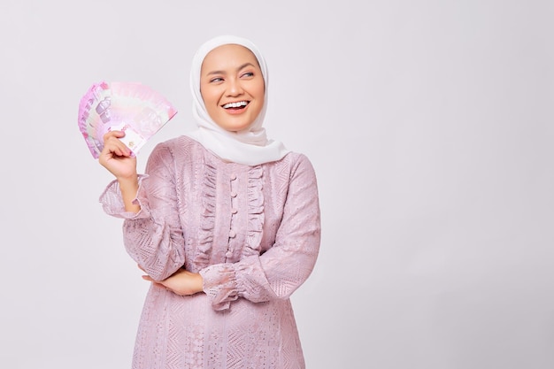 Sorridente bella giovane donna musulmana asiatica che indossa l'hijab e abito viola in possesso di denaro contante rupia banconote mentre guardando da parte isolato su sfondo bianco studio