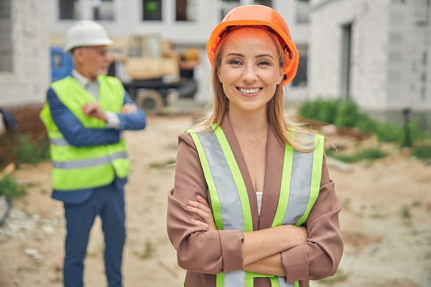 Sorridente bella ingegnere caucasica e un costruttore dietro di lei in piedi in un cantiere edile