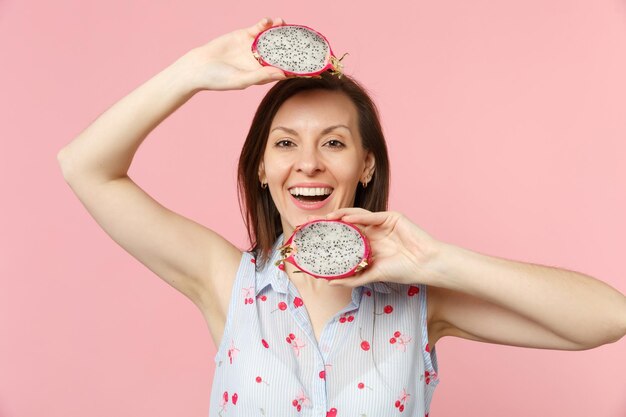 Sorridente bella ragazza in abiti estivi che tiene metà del fresco frutto maturo del drago pitahaya isolato su sfondo rosa muro pastello. Lo stile di vita vivido delle persone si rilassa il concetto di vacanza. Mock up spazio di copia.