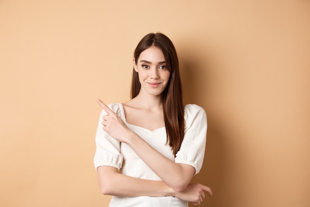 Sorridente bella ragazza in abito bianco rivolto da parte, mostrando il logo a sinistra e fissando la telecamera fiduciosa, in piedi su sfondo beige.