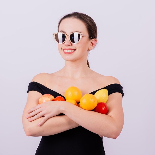 Sorridente bella ragazza in occhiali da sole in costume da bagno nero in posa con frutta e ridendo