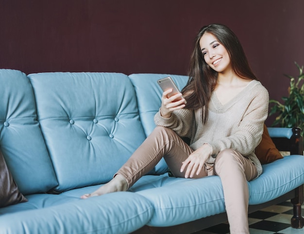 Sorridente bruna giovane donna asiatica in casual utilizzando mobile seduto sul divano a casa