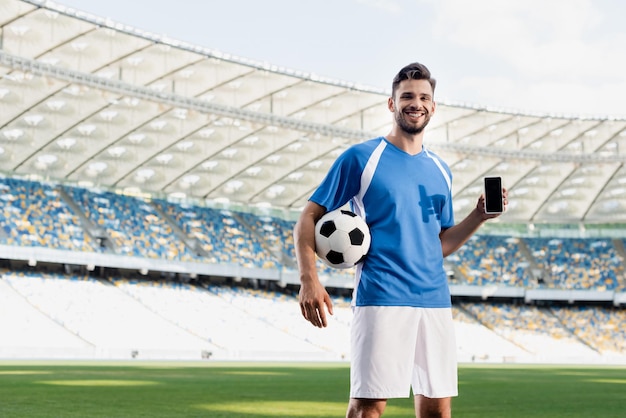 Sorridente calciatore professionista in uniforme blu e bianca con palla che mostra smartphone con vuoto